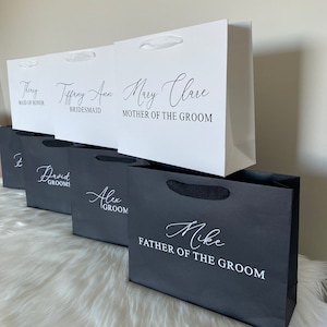 May include: Five white and black gift bags with handles. The bags are labelled with names and titles, such as "Maid of Honour", "Bridesmaid", "Mother of the Groom", "Groom", and "Father of the Groom".