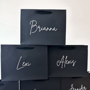 May include: A stack of five black gift bags with white lettering. The names Brianna, Lexi, Alexis, Koryanne, and Jennifer are written on the bags.