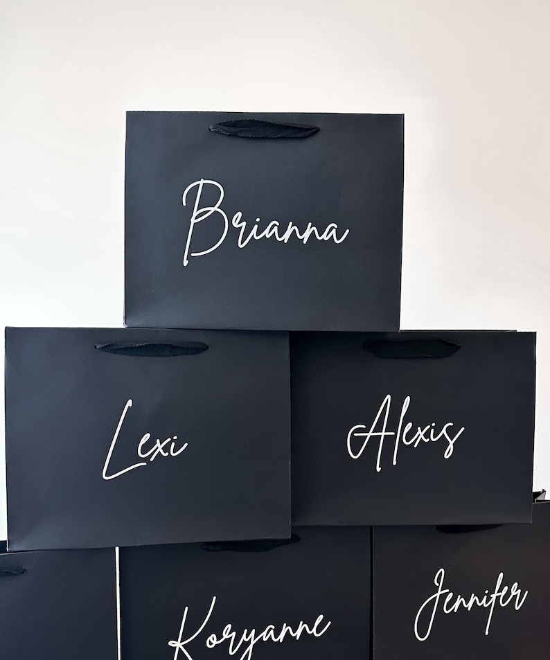 May include: A stack of five black gift bags with white lettering. The names Brianna, Lexi, Alexis, Koryanne, and Jennifer are written on the bags.