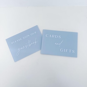 May include: Two rectangular light blue acrylic signs with white lettering. One sign reads "PLEASE SIGN OUR guestbook" and the other reads "CARDS and GIFTS" in a combination of block and script fonts. The signs have a smooth, matte finish.