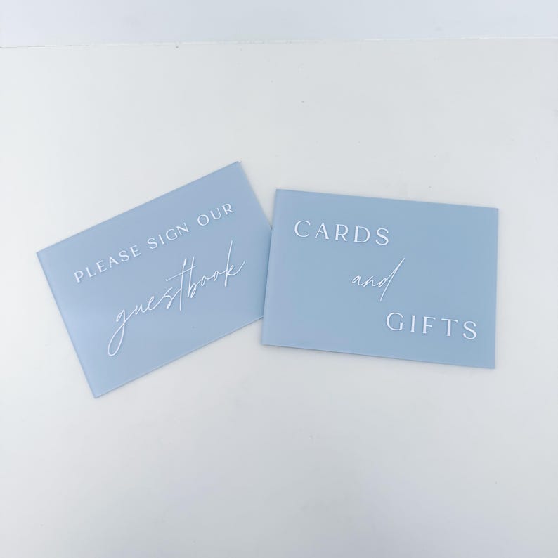 May include: Two rectangular light blue acrylic signs with white lettering. One sign reads "PLEASE SIGN OUR guestbook" and the other reads "CARDS and GIFTS" in a combination of block and script fonts. The signs have a smooth, matte finish.