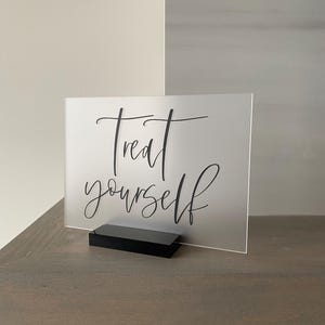 K&ouml;nnte beinhalten: Ein mattes Acrylschild mit einem schwarzen St&auml;nder. Das Schild tr&auml;gt die Aufschrift "Treat yourself" in einer kursiv geschriebenen Schrift.