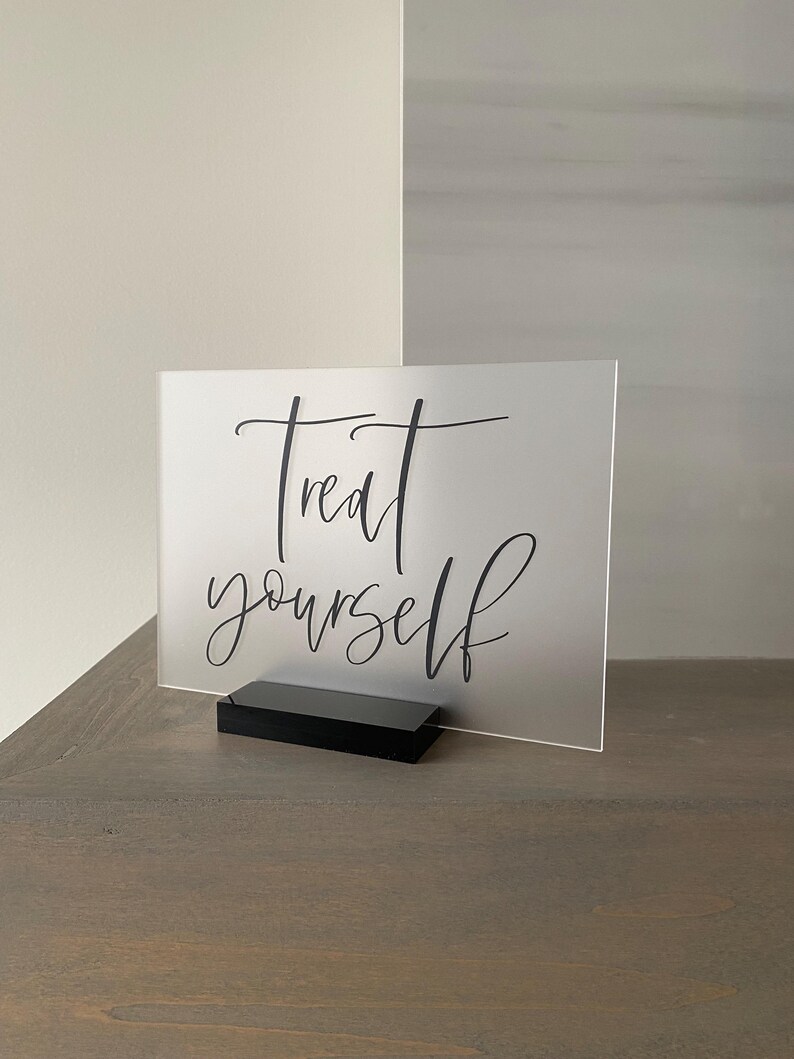 K&ouml;nnte beinhalten: Ein mattes Acrylschild mit einem schwarzen St&auml;nder. Das Schild tr&auml;gt die Aufschrift "Treat yourself" in einer kursiv geschriebenen Schrift.