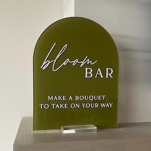 May include: A green arch-shaped sign with white text that reads "bloom BAR" and "MAKE A BOUQUET TO TAKE ON YOUR WAY". The sign is on a clear acrylic stand.
