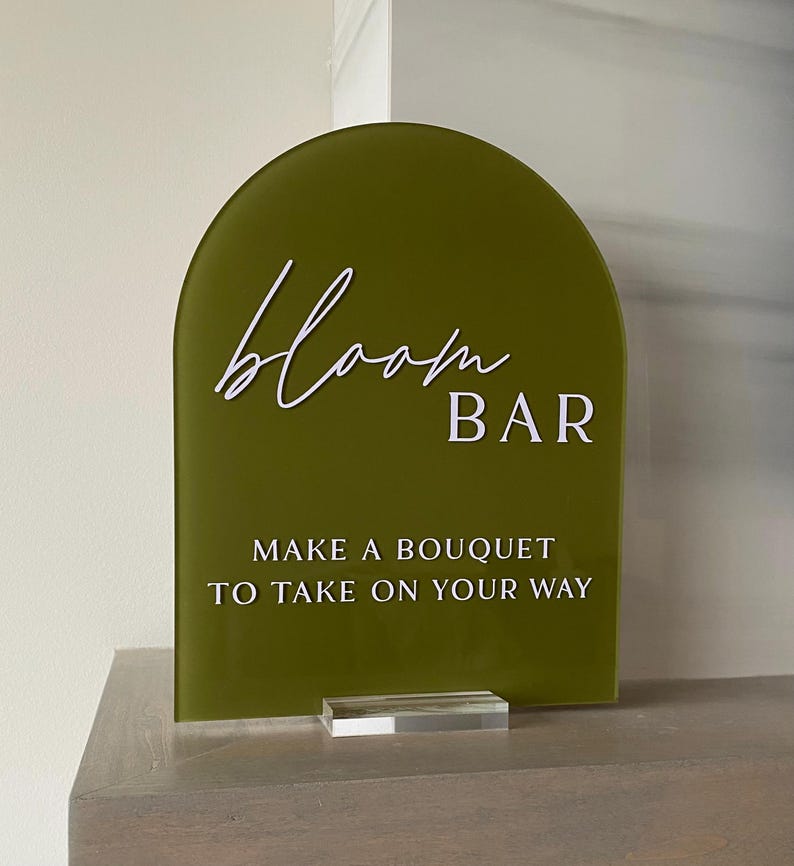 May include: A green arch-shaped sign with white text that reads "bloom BAR" and "MAKE A BOUQUET TO TAKE ON YOUR WAY". The sign is on a clear acrylic stand.