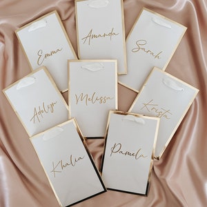 May include: Seven white place cards with gold trim and gold calligraphy names: Emma, Amanda, Sarah, Adelyn, Melissa, Krista, and Kalia. The cards are arranged on a light pink satin fabric.