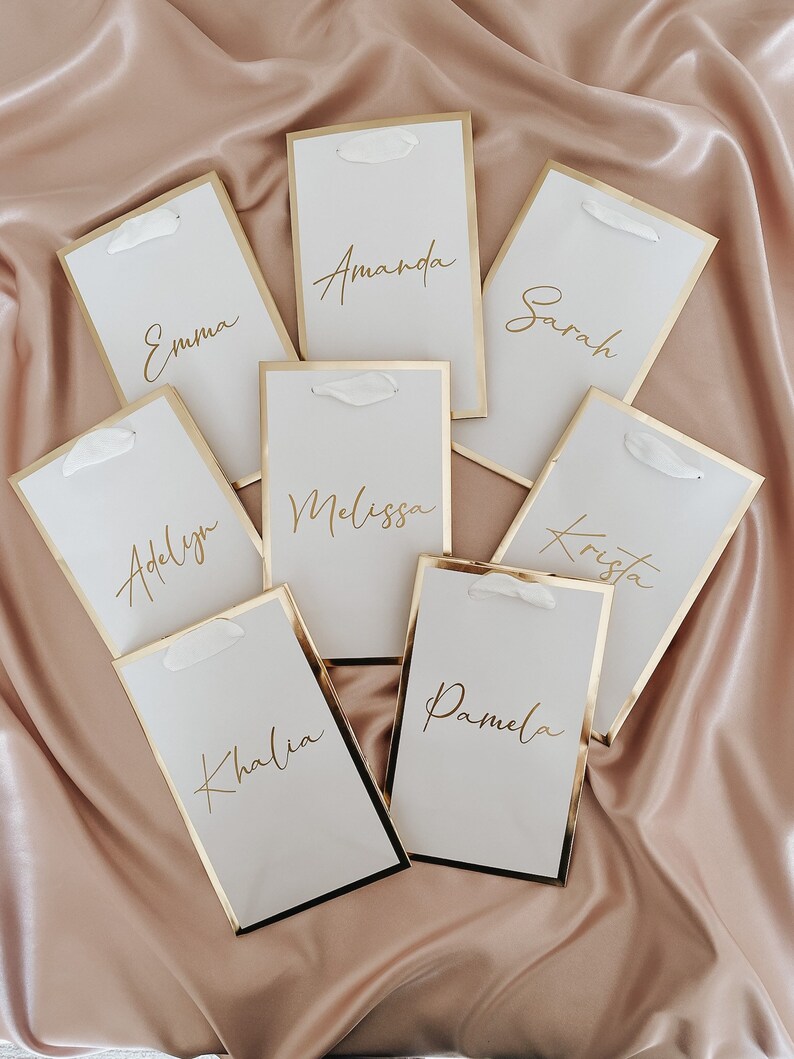 May include: Seven white place cards with gold trim and gold calligraphy names: Emma, Amanda, Sarah, Adelyn, Melissa, Krista, and Kalia. The cards are arranged on a light pink satin fabric.