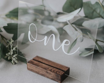 May include: A clear acrylic table number with the word "one" written in white calligraphy. The number is displayed on a wooden stand.