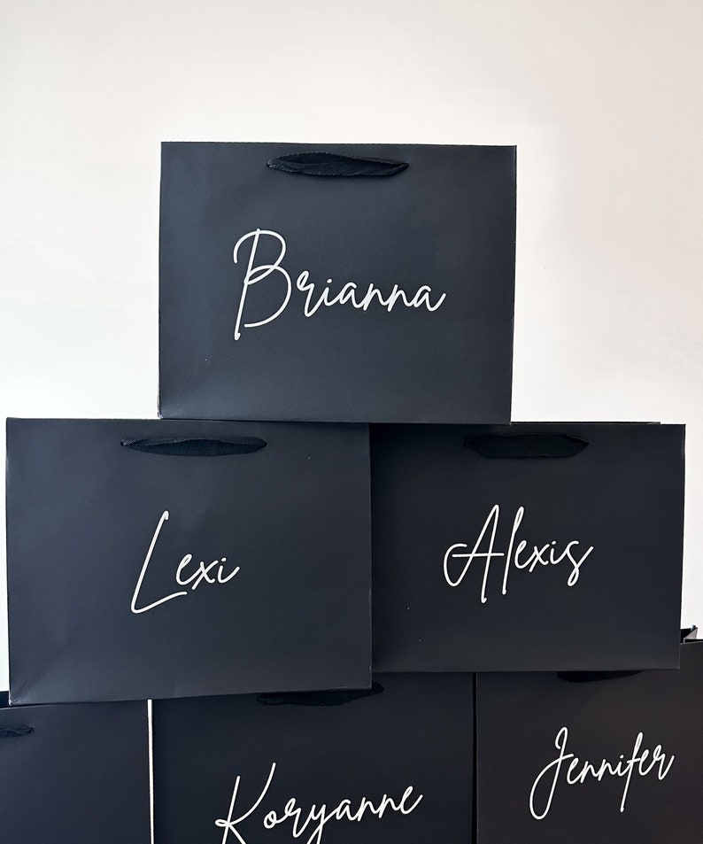 May include: Stack of five black gift bags with white lettering. The names Brianna, Lexi, Alexis, Koryanne, and Jennifer are written on the bags.