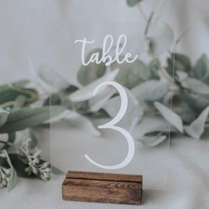 May include: Clear acrylic table number with the text "table 3" in white lettering, on a wooden base.