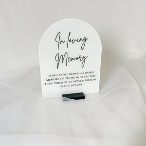 以下が含まれることがあります： 「In loving memory」というテキストが書かれた白いアクリル製のアーチと、その下に「This candle burns in loving memory of those who are not here today but forever present in our hearts.」というテキストが書かれた小さなテキストブロックがあります。