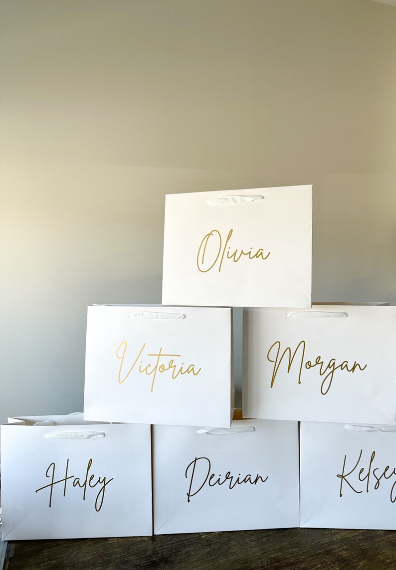 May include: Six white gift bags with gold lettering. The names Olivia, Victoria, Morgan, Haley, Deirion, and Kelsey are written on the bags.