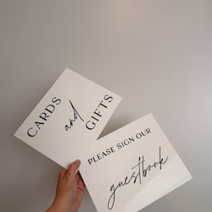 May include: Two rectangular white acrylic signs with black lettering. One sign features the text CARDS and GIFTS with the word and in script. The second sign features the text PLEASE SIGN OUR guestbook with the word guestbook in script.