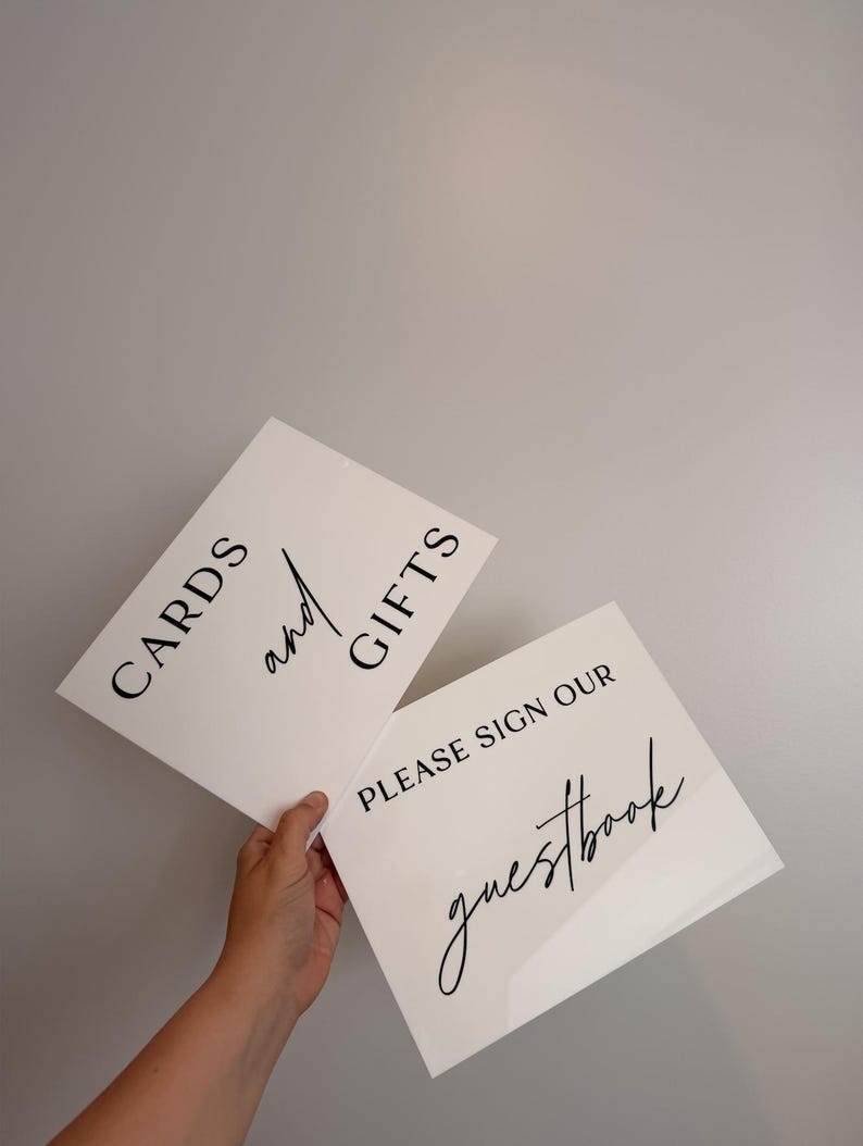 May include: Two rectangular white acrylic signs with black lettering. One sign features the text CARDS and GIFTS with the word and in script. The second sign features the text PLEASE SIGN OUR guestbook with the word guestbook in script.