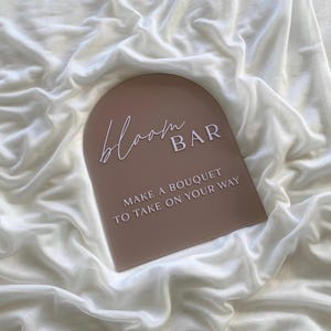 May include: A light brown arch-shaped sign with the text "bloom BAR" in white script font. The sign also has the text "MAKE A BOUQUET TO TAKE ON YOUR WAY" in white.
