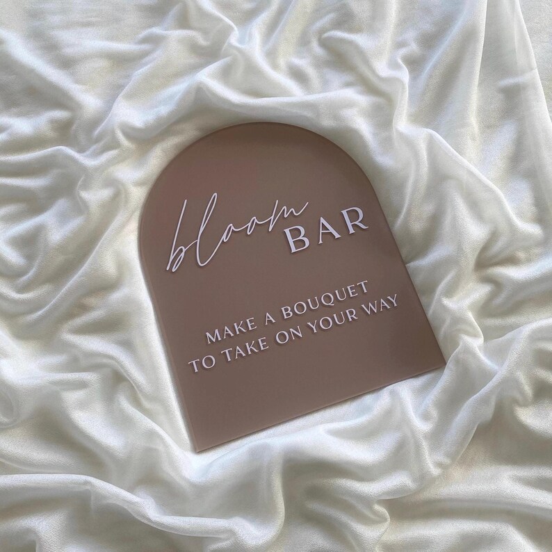 May include: A light brown arch-shaped sign with the text "bloom BAR" in white script font. The sign also has the text "MAKE A BOUQUET TO TAKE ON YOUR WAY" in white.