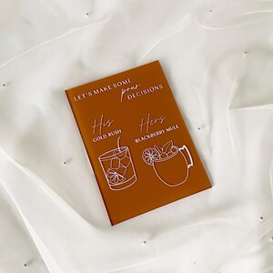May include: A brown acrylic sign with white text that reads "Let's make some decisions". The sign has two drink options, "His" with a gold rush cocktail and "Hers" with a blackberry mule cocktail.