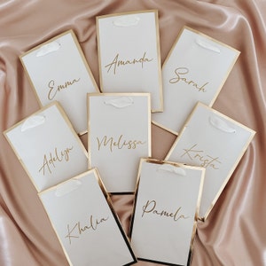 May include: Eight white place cards with gold trim and gold calligraphy names: Emma, Amanda, Sarah, Adelyn, Melissa, Krista, Kalia, and Pamela.