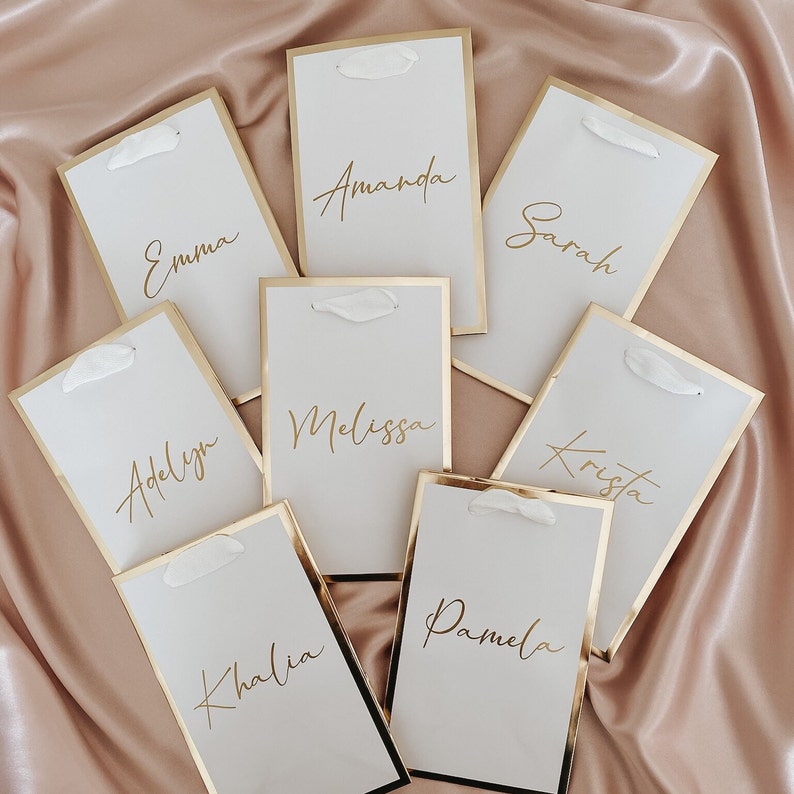 May include: Eight white place cards with gold trim and gold calligraphy names: Emma, Amanda, Sarah, Adelyn, Melissa, Krista, Kalia, and Pamela.