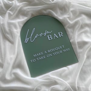 May include: A green arch-shaped sign with white text that reads "bloom BAR" and "MAKE A BOUQUET TO TAKE ON YOUR WAY".