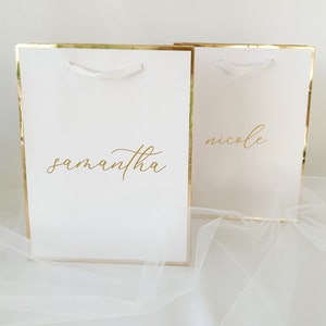 May include: Two white gift bags with gold trim and white ribbon handles. The bags are personalized with the names "samantha" and "nicole" in gold lettering.