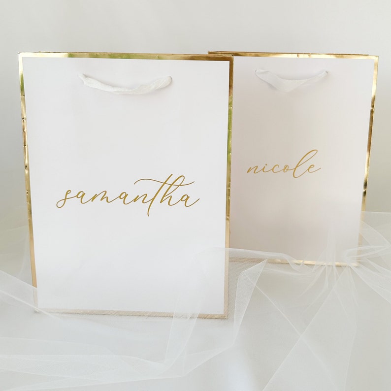May include: Two white gift bags with gold trim and white ribbon handles. The bags are personalized with the names "samantha" and "nicole" in gold lettering.