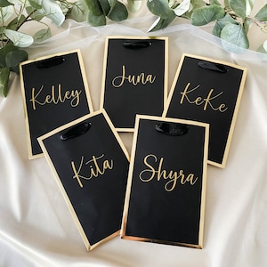 May include: Five black gift bags with gold trim and black ribbon handles. Each bag has a name written in gold lettering: Kelley, Kita, June, Keke, and Shyra.