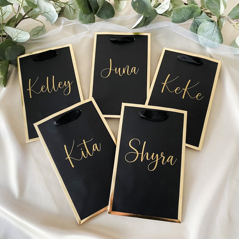 May include: Five black gift bags with gold trim and black ribbon handles. Each bag has a name written in gold lettering: Kelley, Kita, June, Keke, and Shyra.