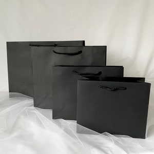May include: Four black paper gift bags of different sizes, arranged in a row from largest to smallest. The bags have black rope handles.
