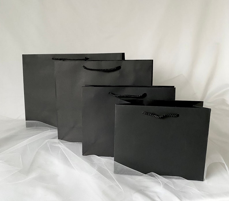 May include: Four black paper gift bags of different sizes, arranged in a row from largest to smallest. The bags have black rope handles.