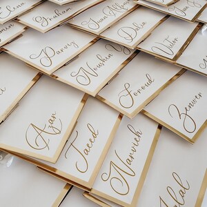 May include: A close-up of a stack of white place cards with gold trim. Each card has a name written in elegant calligraphy. The names include Azan, Noushin, Delia, Foroud, Narvich, and Jalal.