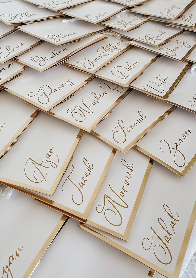 May include: A close-up of a stack of white place cards with gold trim. Each card has a name written in elegant calligraphy. The names include Azan, Noushin, Delia, Foroud, Narvich, and Jalal.