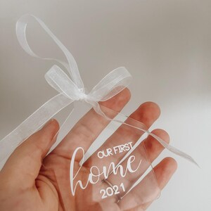 Puede incluir: Un adorno transparente de acr&iacute;lico con una cinta blanca. El adorno tiene el texto "Our First Home 2021" escrito en &eacute;l.