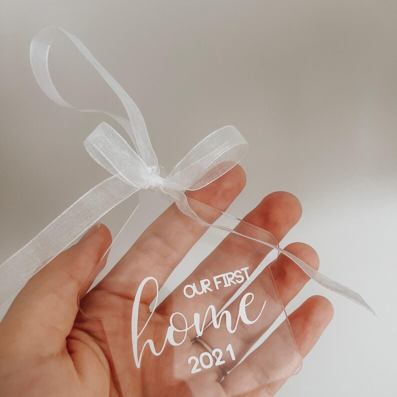Puede incluir: Un adorno transparente de acr&iacute;lico con una cinta blanca. El adorno tiene el texto "Our First Home 2021" escrito en &eacute;l.