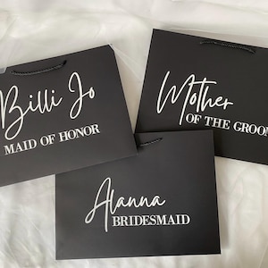 Op de afbeelding: Drie zwarte cadeauzakken met witte tekst. De tassen hebben de tekst "Billi Jo Maid of Honor", "Mother of the Groom" en "Alanna Bridesmaid".