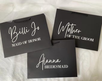 以下が含まれることがあります： 白い文字が書かれた黒いギフトバッグが3つ。バッグには「Billi Jo Maid of Honor」、「Mother of the Groom」、「Alanna Bridesmaid」と書いてあります。