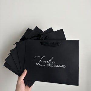 May include: A stack of black gift bags with white text that reads "Linda Bridesmaid".