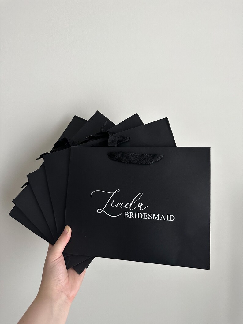 May include: A stack of black gift bags with white text that reads "Linda Bridesmaid".