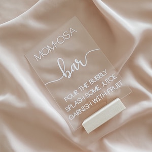 May include: Clear acrylic sign with white text that reads "MOM-OSA bar POUR THE BUBBLY SPLASH SOME JUICE GARNISH WITH FRUIT". The sign is propped up on a small wooden block.