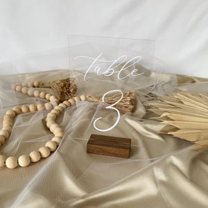 May include: Clear acrylic table number sign with the text "Table 3" in white calligraphy. The sign is on a wooden base.