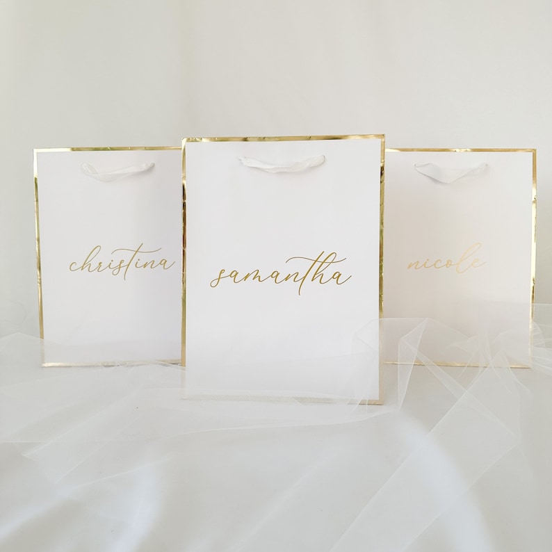 May include: Three white gift bags with gold trim and white ribbon handles. The bags are personalized with the names "Christina", "Samantha", and "Nicole" in gold lettering.