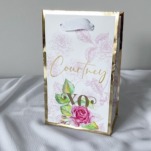 May include: A white gift bag with a gold border and floral design. The bag has a white ribbon handle and the name "Courtney" and "XO" written in gold lettering.