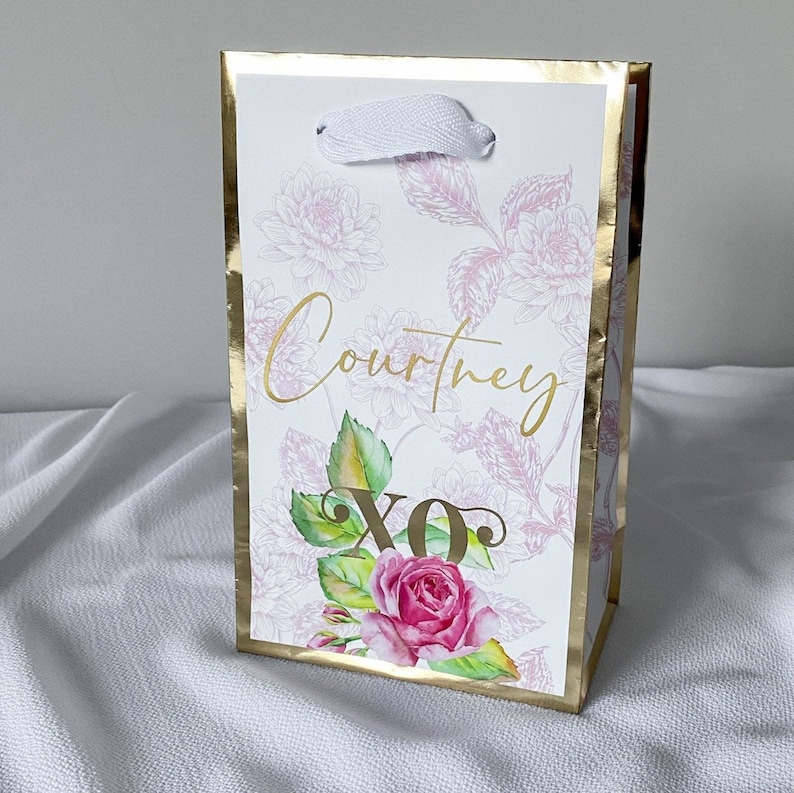 May include: A white gift bag with a gold border and floral design. The bag has a white ribbon handle and the name "Courtney" and "XO" written in gold lettering.