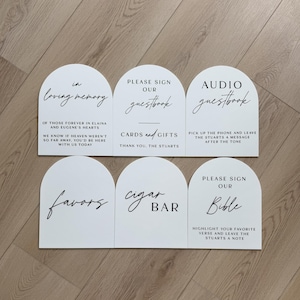 May include: Six white arched signs with black text. The signs feature various wedding-related labels including In Loving Memory, Please Sign Our Guestbook, Cards and Gifts, Audio Guestbook, Favors, Cigar Bar, and Please Sign Our Bible with custom text.