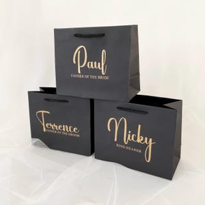May include: Three black gift bags with gold lettering. The bags are stacked on top of each other. The top bag says "Paul, Father of the Bride". The middle bag says "Terrence, Father of the Groom". The bottom bag says "Nicky, Ring Bearer".