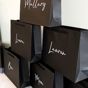 May include: A stack of black gift bags with white lettering. The names on the bags are Mallory, Laura, Lauren, Ben, and Mom.