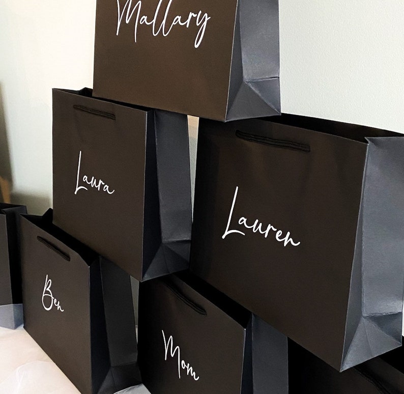 May include: A stack of black gift bags with white lettering. The names on the bags are Mallory, Laura, Lauren, Ben, and Mom.