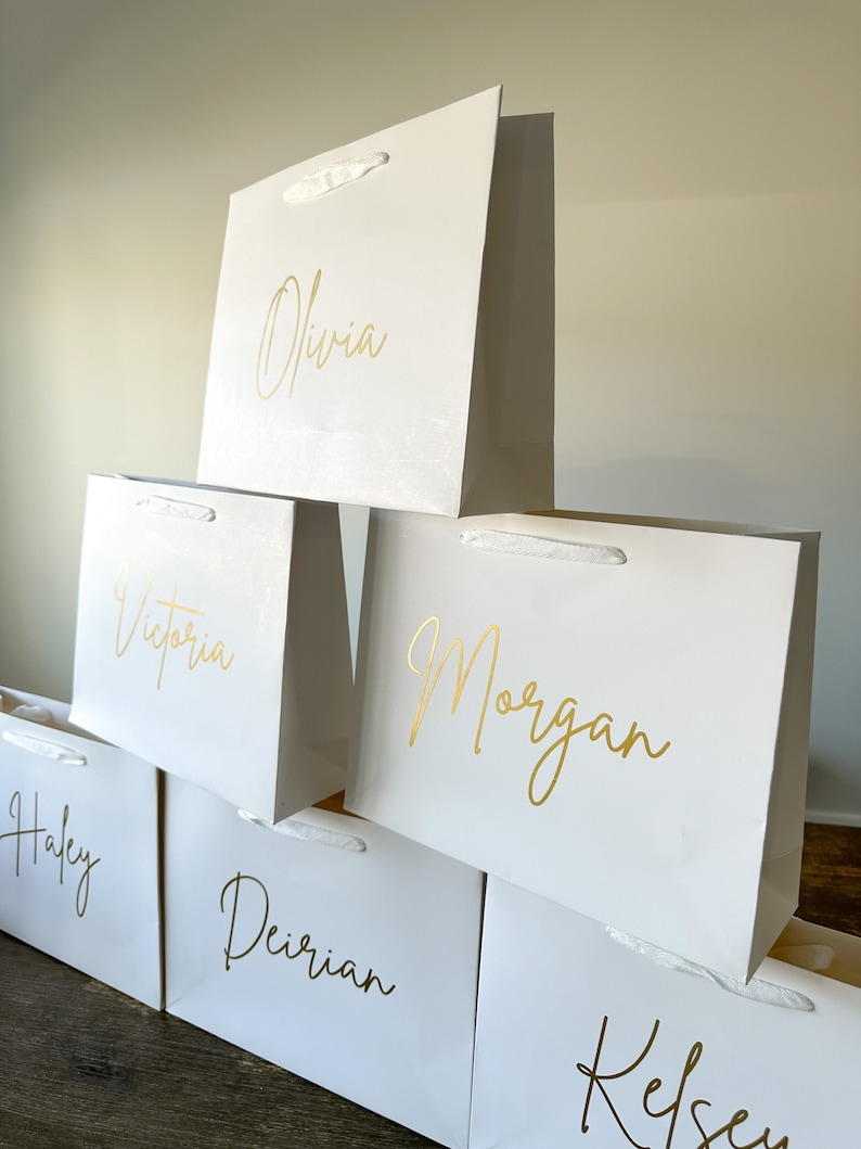 May include: Stack of six white gift bags with gold lettering. The names Olivia, Victoria, Morgan, Haley, Deirien, and Kelsey are written on the bags.