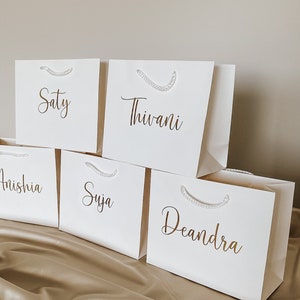 May include: Five white gift bags with gold lettering. The names on the bags are Saty, Thivani, Anishia, Suja, and Deandra.