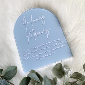 以下が含まれることがあります： 「In loving memory」というテキストと、出席していない人を偲ぶメッセージが書かれた薄い青色のアーチ型の看板。看板は白いふわふわの表面の上に置かれています。